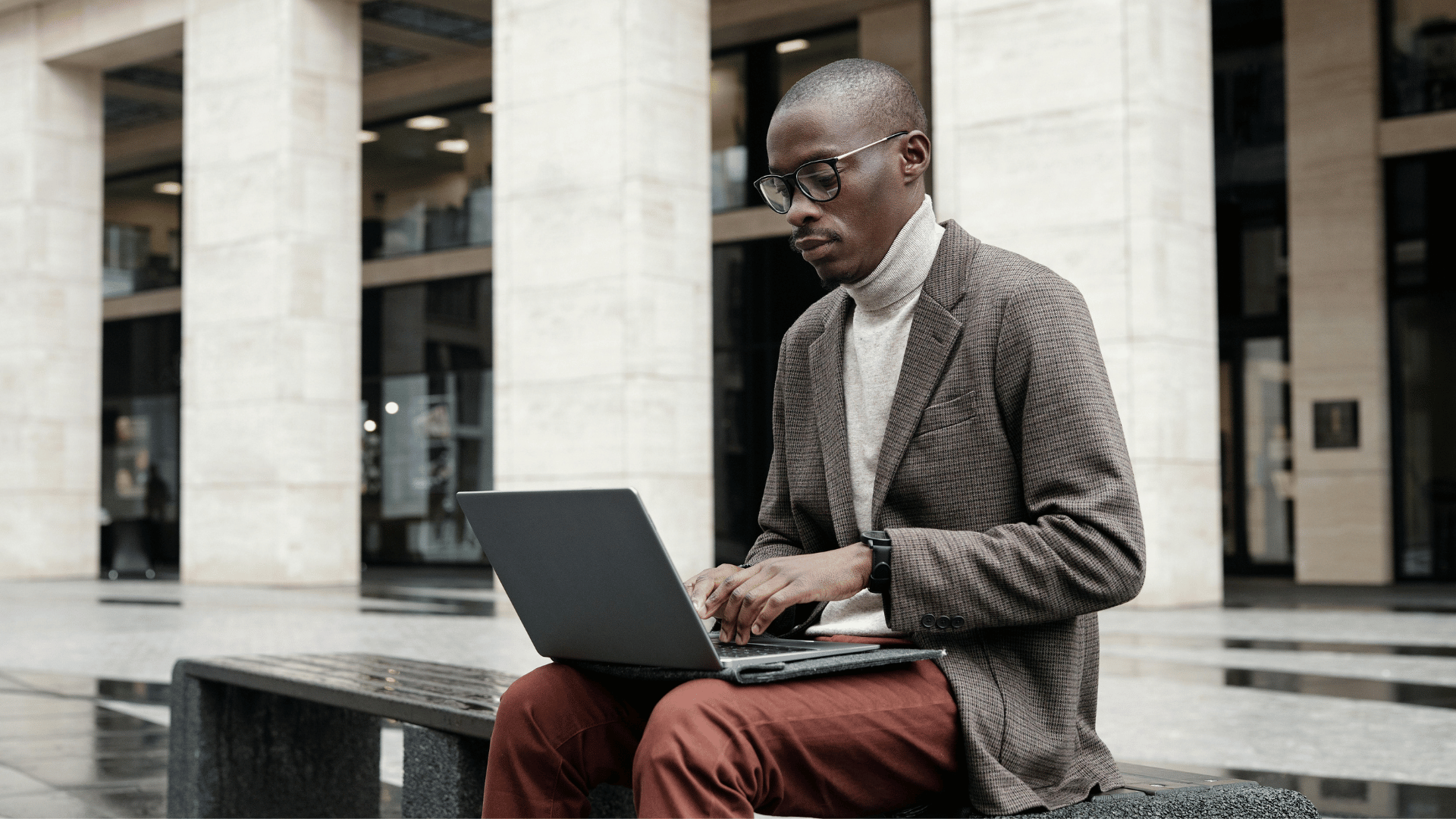 An African American man sits on a bench with his laptop open on his lap.